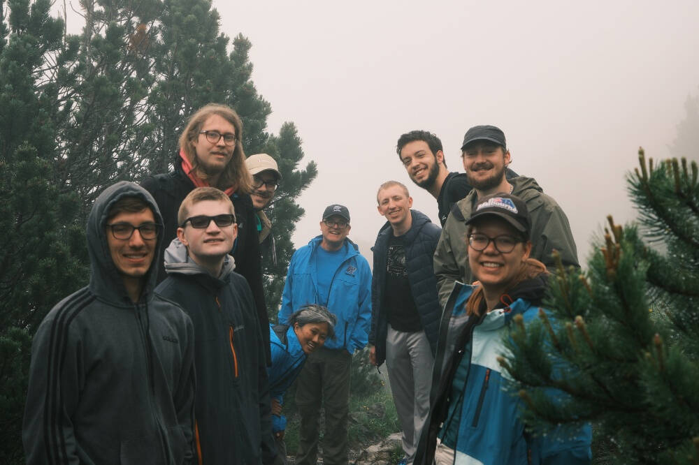 A group of eleven people dressed in outdoor and hiking gear pose together in a misty forest surrounded by pine trees. Most are smiling and appear to be enjoying the hike. The atmosphere is foggy, creating a serene and adventurous setting.
